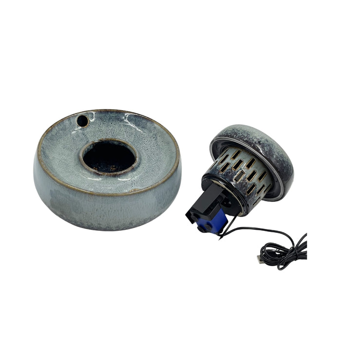 A ceramic ring-shaped vessel with a glossy speckled gray and blue glaze, shown alongside a small black and blue mechanical component with a black wire on a plain white background.