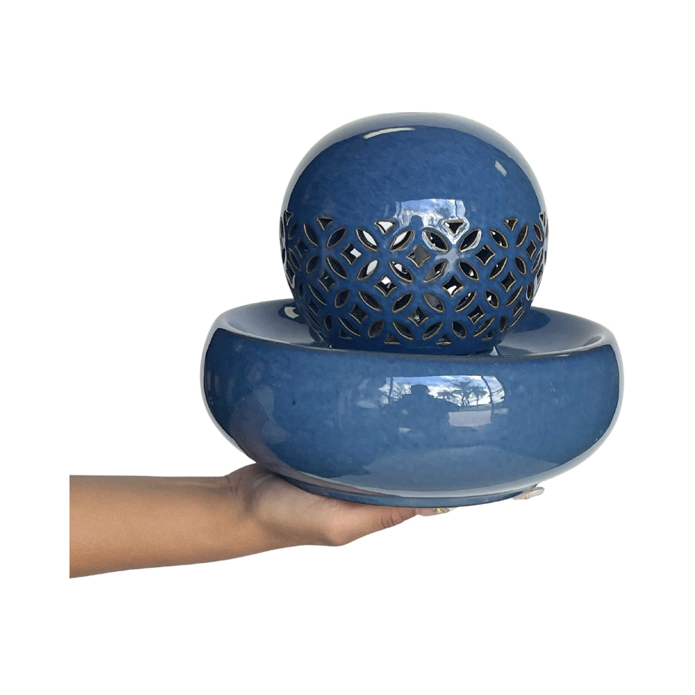 A glossy blue ceramic water feature consisting of a round basin and a perforated decorative sphere, shown being held by a person's hand against a white background.