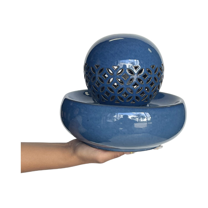 A glossy blue ceramic water feature consisting of a round basin and a perforated decorative sphere, shown being held by a person's hand against a white background.