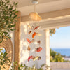 A hanging shell wind chime with multiple natural seashells on strings, suspended from a ceiling outdoors on a porch with a blurred sea and plants in the background.