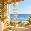 A boho-style seashell wind chime hanging from a wooden porch railing, with a blurred background of the blue ocean and sky.