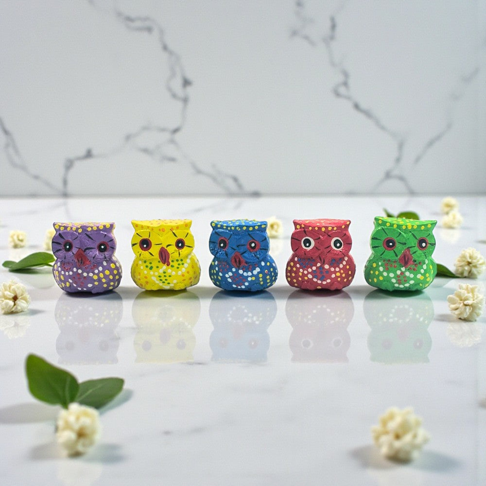 Five colorful owl figurines on a marble surface with flowers.