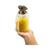 Hand holding a yellow candle in a glass jar with a brown drawstring bag on a white background