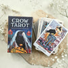 Crow Tarot card set with deck and box on a light surface with flowers.