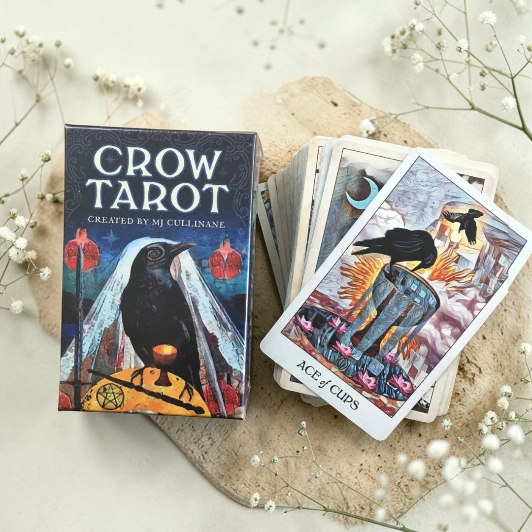 Crow Tarot card set with deck and box on a light surface with flowers.