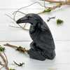 Black stone carving of a raven on a white wooden surface with branches and greenery.