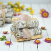 Stack of smudge sticks with rose quartz on a wooden stand, surrounded by flowers on a white wooden surface.